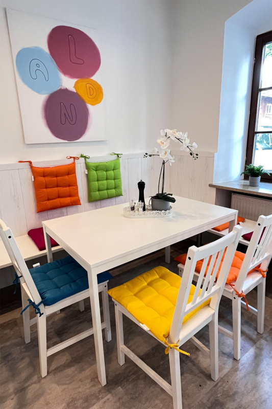 Kinderland Bildungsinstitut Impulskraft Mensa. Gemütliche Sitzecke mit weissem Tisch und Stühlen mit bunten Polstern und einer Orchidee auf dem Tisch. An der Wand hängt ein Bild mit 4 bunten Kreisen und den Buchstaben L A N D